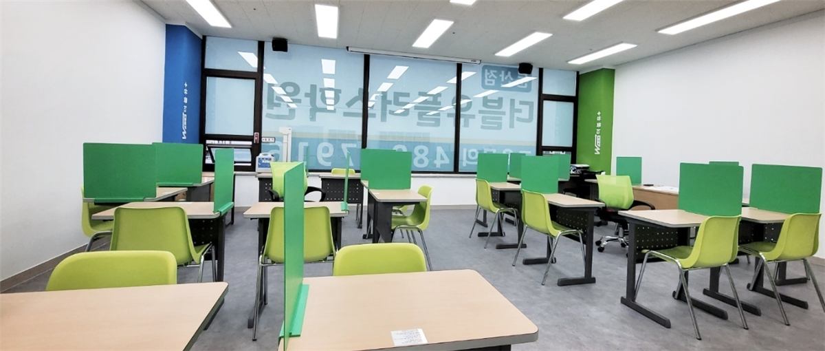 둔산점 수학전문관 교실 — 창가를 따라 초록 가림판 책상이 놓여 있고, 유리창에 더블유플러스학원 둔산점 글자가 비치는 모습