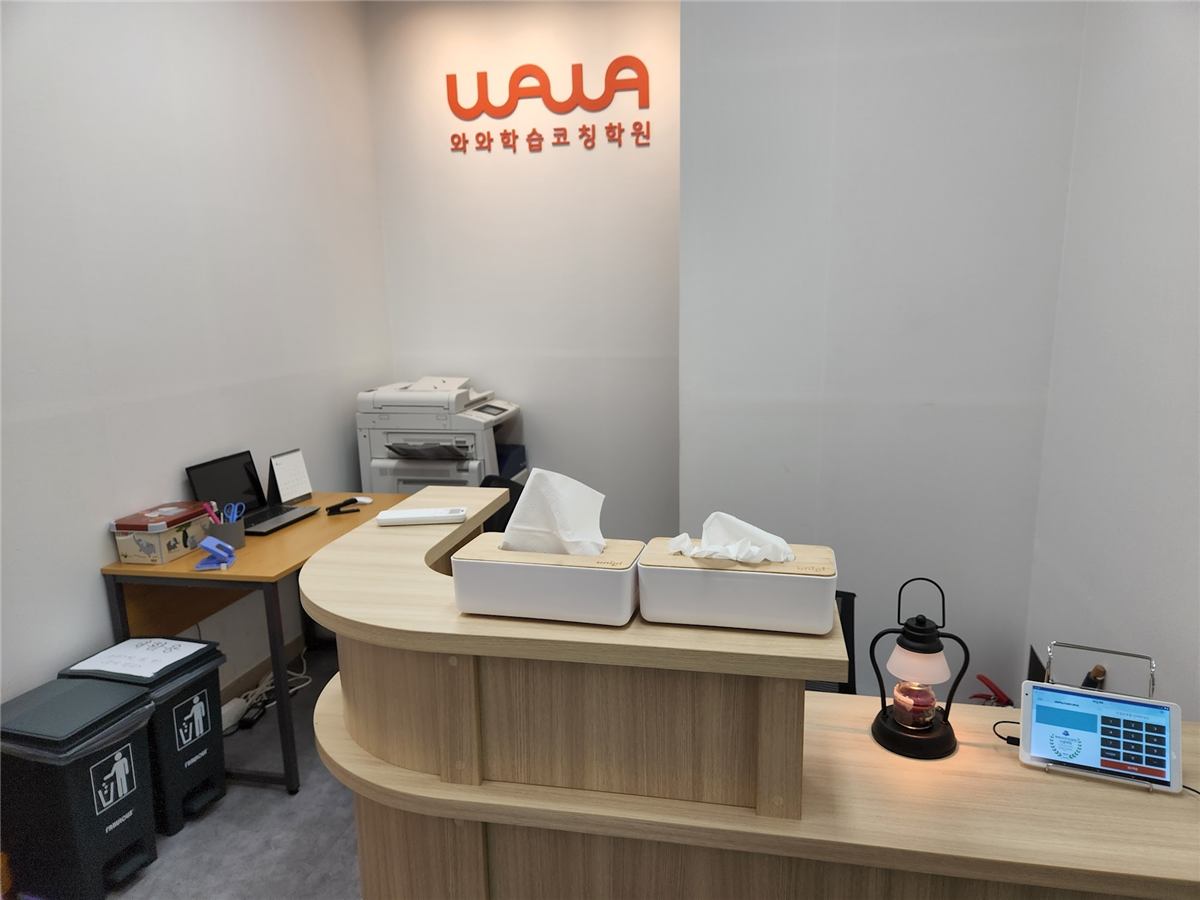 범박점 안내 데스크 — 흰 벽 위쪽에 주황색 WAWA 로고와 와와학습코칭학원 글자가 붙어 있고, 그 아래 원목 곡선 데스크가 놓여 있다. 데스크 위에 티슈 상자 두 개와 스탠드 조명, 출결 확인용 태블릿이 있고, 뒤쪽으로 복합기와 노트북이 놓인 업무용 책상이 보인다