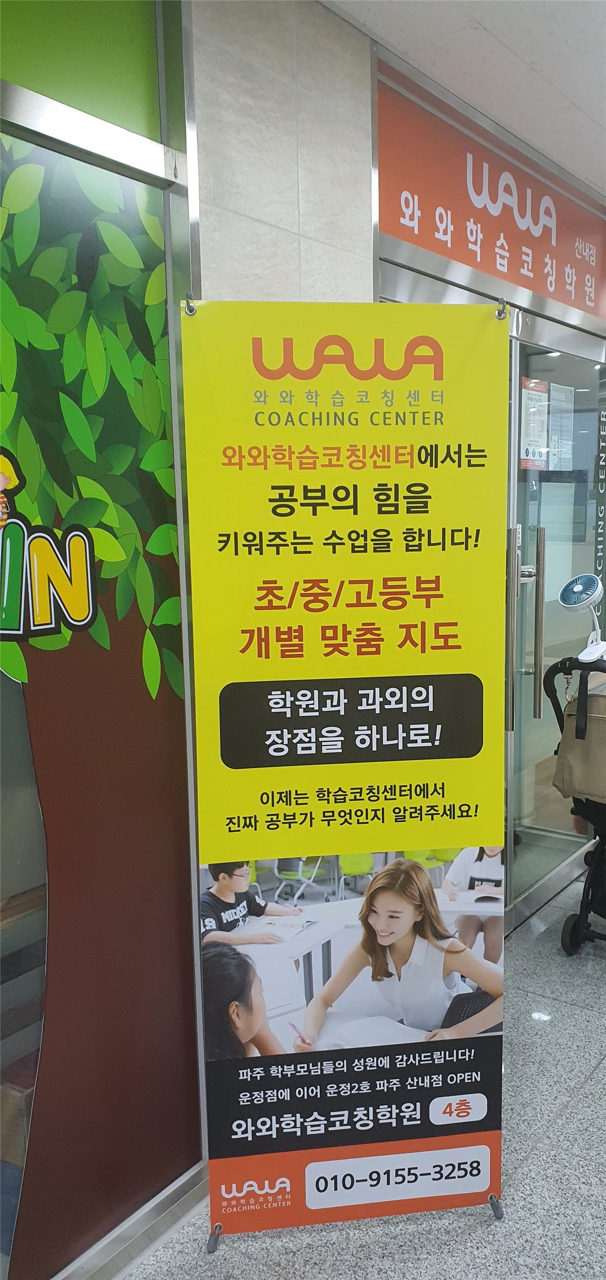산내점 입구 앞에 세워진 안내 배너 — 노란 바탕에 와와학습코칭센터 로고와 '공부의 힘을 키워주는 수업을 합니다', '초/중/고등부 개별 맞춤 지도'라는 문구가 적혀 있다. 배너 아래에는 '운정점에 이어 운정2호 파주 산내점 OPEN, 와와학습코칭학원 4층'이라고 쓰여 있고, 뒤쪽 유리문 위에 주황색 산내점 간판이 보인다