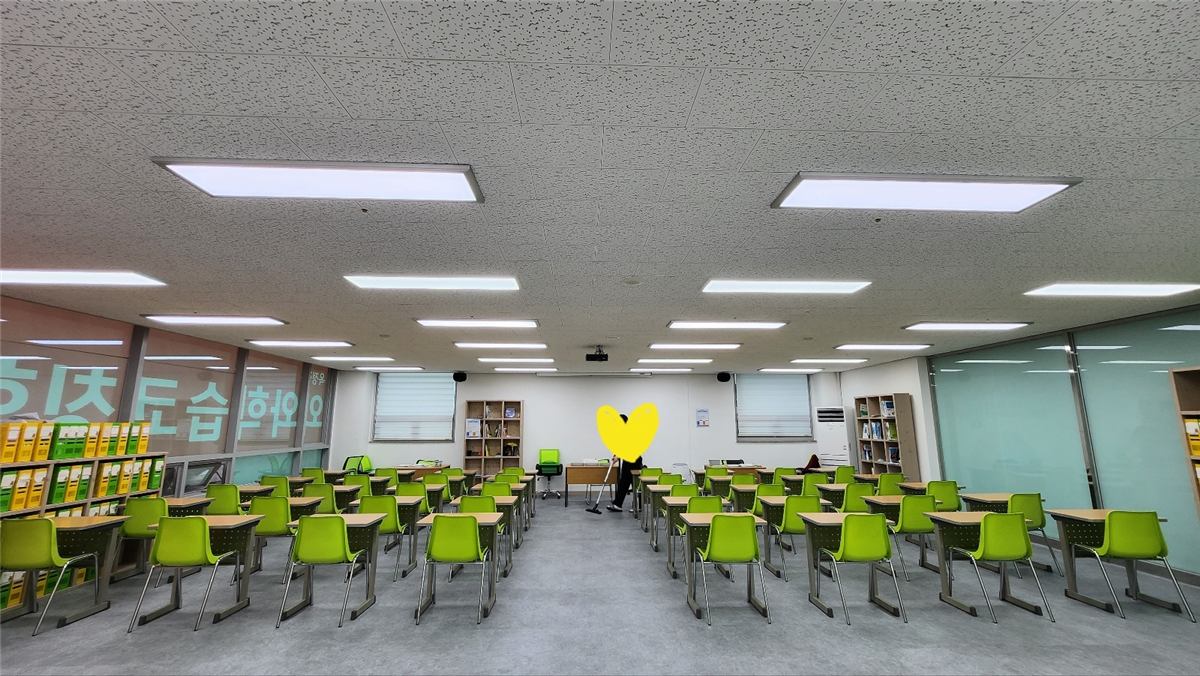 옥정점 넓은 학습실 전경 — 연두색 의자와 책상이 여러 줄로 가지런히 놓여 있고 천장에는 긴 조명이 이어져 있다. 왼쪽 벽면에는 파일박스가 꽂힌 수납장과 분홍색 브랜드 월이, 안쪽에는 책이 꽂힌 책장 두 개가 보인다