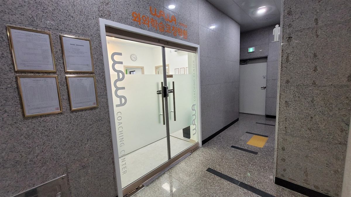 전주혁신점 입구 — 건물 복도 안쪽에 있는 유리 양문 위에 주황색 와와 로고와 와와학습코칭학원 글자가 붙어 있고, 유리문에는 WAWA COACHING CENTER가 세로로 인쇄되어 있다. 왼쪽 화강암 벽에는 학원 등록 관련 안내문이 액자에 담겨 걸려 있다