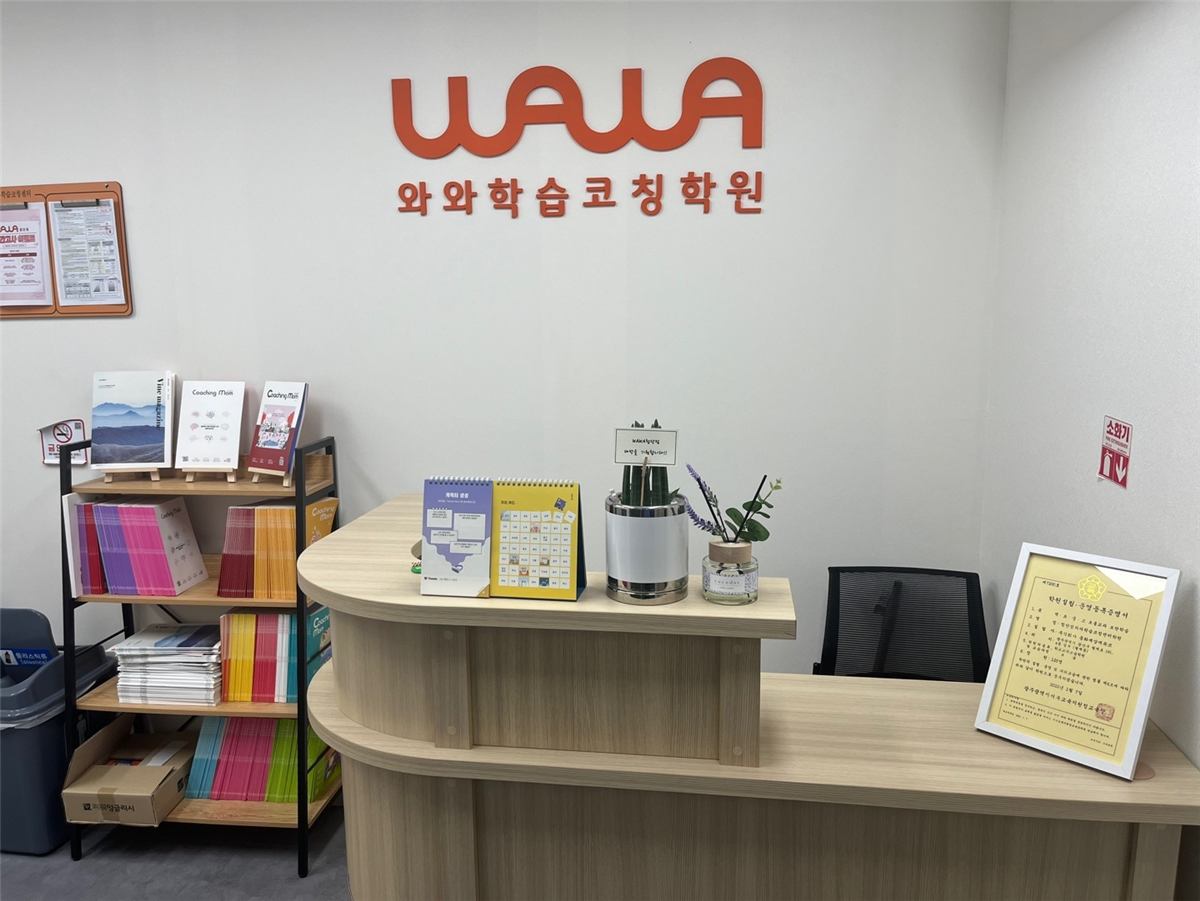 첨단점 안내 데스크 — 흰 벽의 주황색 WAWA 와와학습코칭학원 로고, 나무색 곡선 데스크 위 탁상 달력과 화분, 액자에 넣은 학원 설립·운영 등록증명서, 왼쪽 선반에 꽂힌 Coaching Mom 학부모 책자와 교재